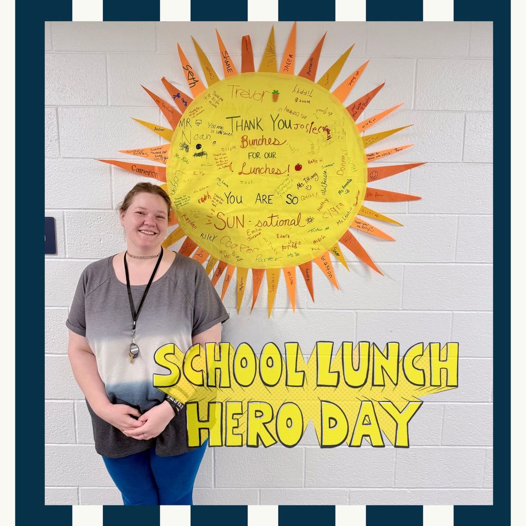 Alex smiles next to a large paper sunshine on the wall. It says "Thank you bunches for our lunches." It is covered with messages and signatures from staff and students.  Texts reads : School Lunch Hero Day."