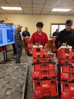 Several bright red toolboxes stacked on a table in the foreground; multiple people stand behind the table in a room with carpeted flooring, while a large display screen and an exit sign are visible in the background.