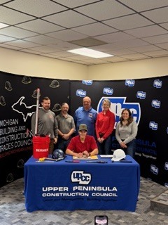 Five people seated and standing behind a blue table with a sign reading “Upper Peninsula” at an indoor event; backdrop includes logos and a black wall graphic, with a white hard hat placed on the table.