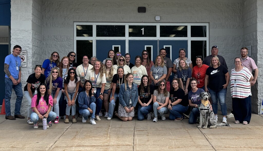 A group of smiling school staff pose in front of the entrance to the school.