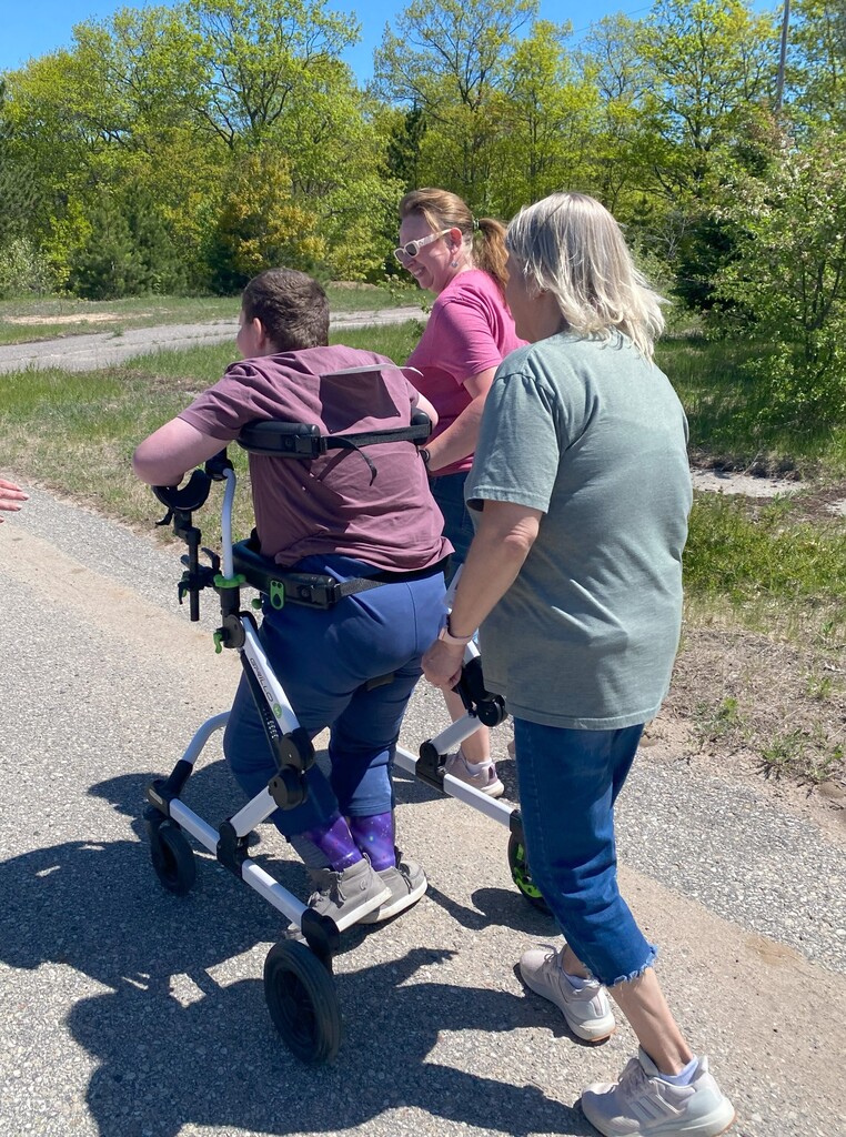 Two smiling staff walk with a student utilizing a gait trainer. There are trees and sunshine in the background. 
