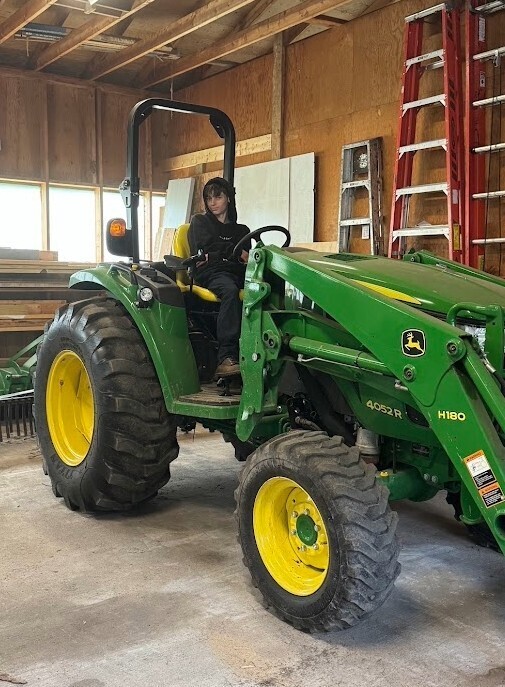 A young man sits in the driver seat of a bright green and yellow John Deere tractor.