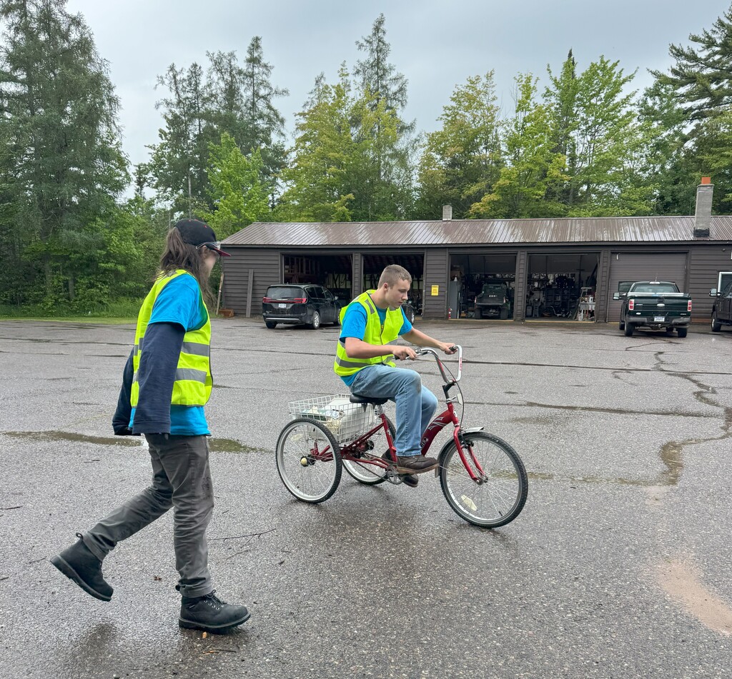 A young man in a bright yellow safety vest rides a 3 wheel bike with a large basket in the back. Another yound man in a safety vest stands nearby.