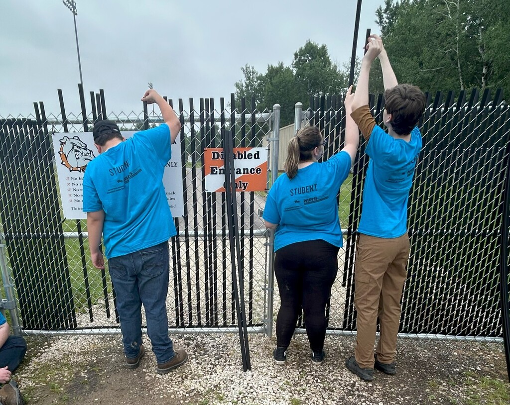 Three students in bright blue Summer Youth and Learn t shirts face away from the camera; feeding black slats into a chain link fence.
