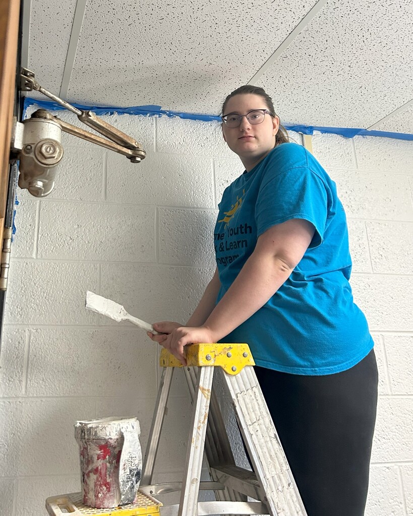 A student stands on a ladder with paint and a paint brush.
