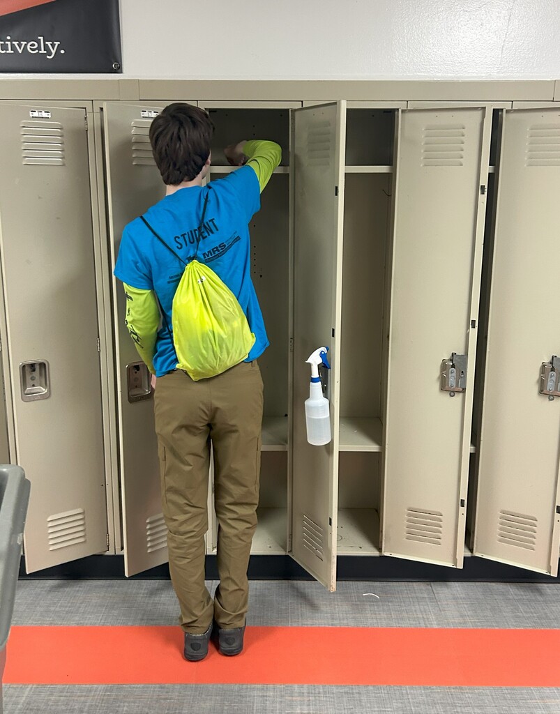 A student stands by a row of lockers. He is wiping down the inside shelf with cleaner.