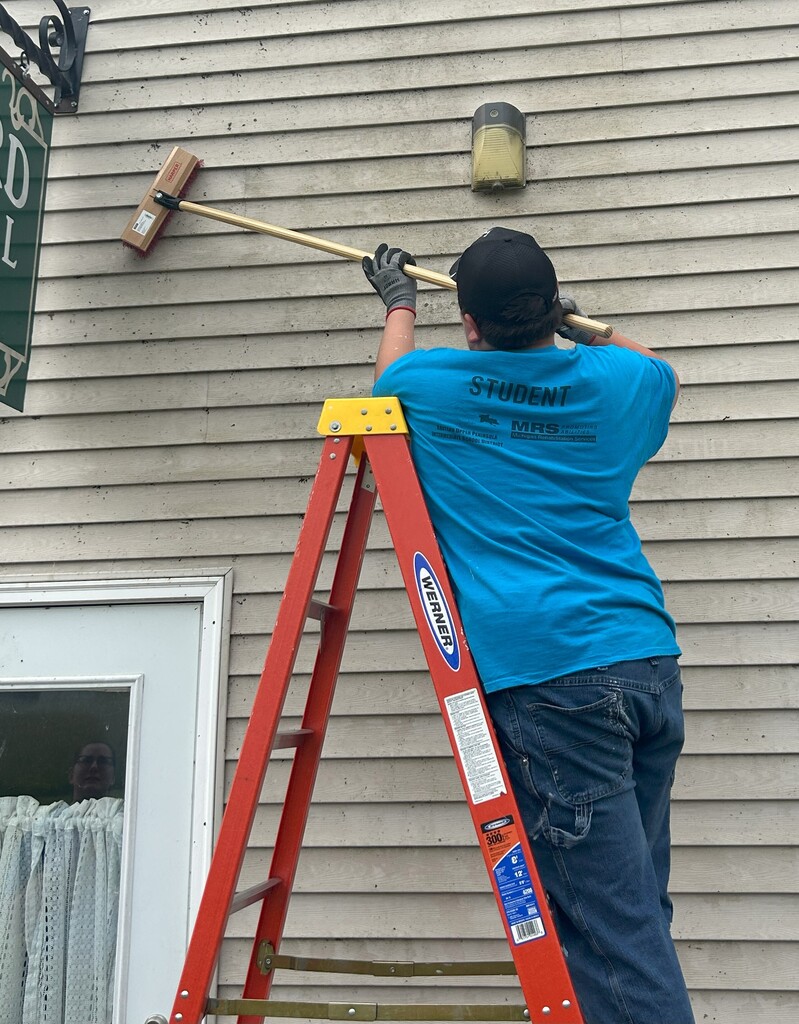 A student on a ladder uses a long brush to clean siding on a building.
