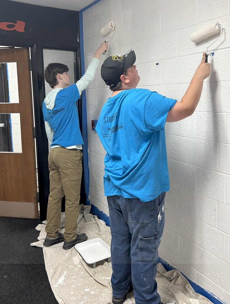 Two students face a wall painting with paint rollers.
