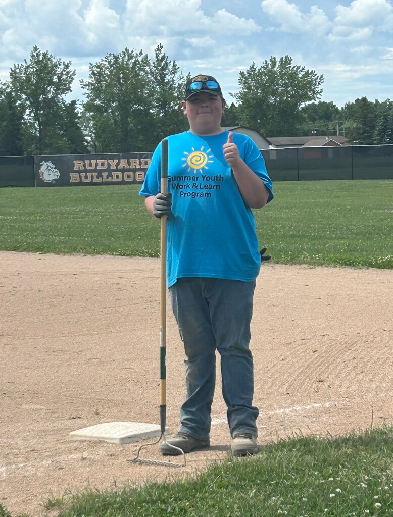A student standing on a baseball field with a rake gives a thumbs up. A sign in the background says "Rudyard Bulldogs."