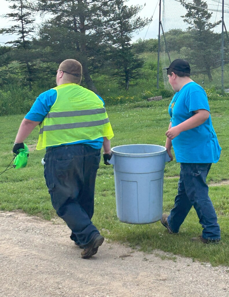 Two students walk away from the camera, carrying a garbage can in between them.