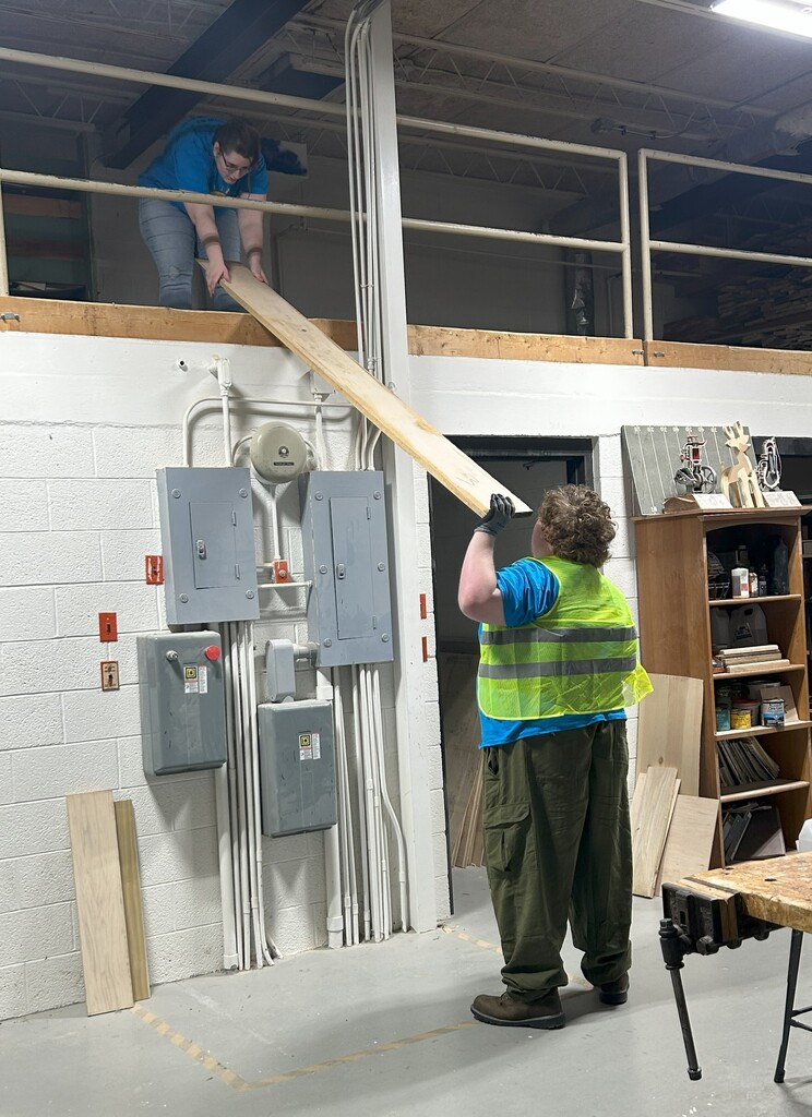 A student feeds a 2x4 wood plank thorugha railing to another student on a higher level.