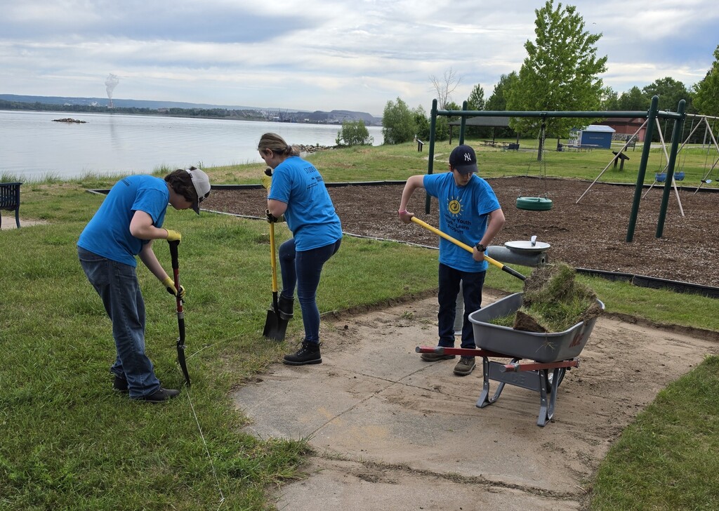 Students are using edging tools to remove overgrown grass on a sidewalk in a park.