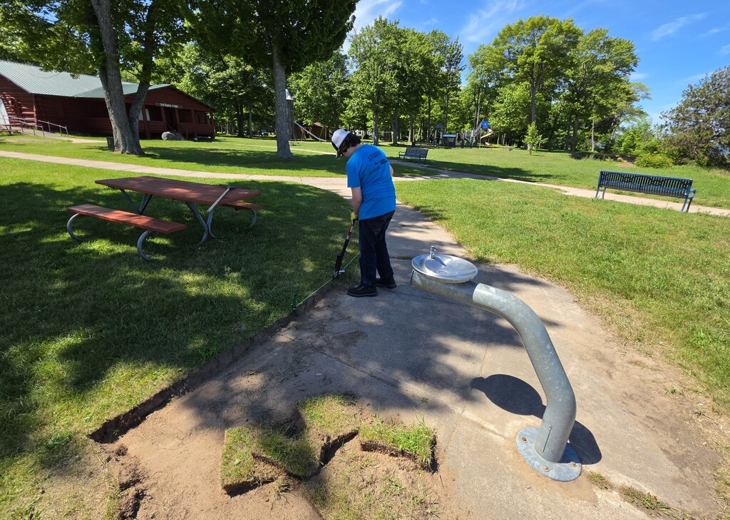 A student is using an edger to remove overgrown grass on a sidewalk in a park.