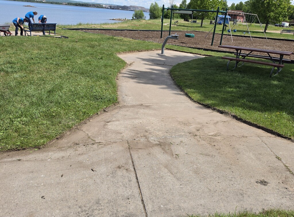 A drinking fountain on a concrete pad in a sunny park. The outside of the pad have recently been edged. to remove overgrowth.
