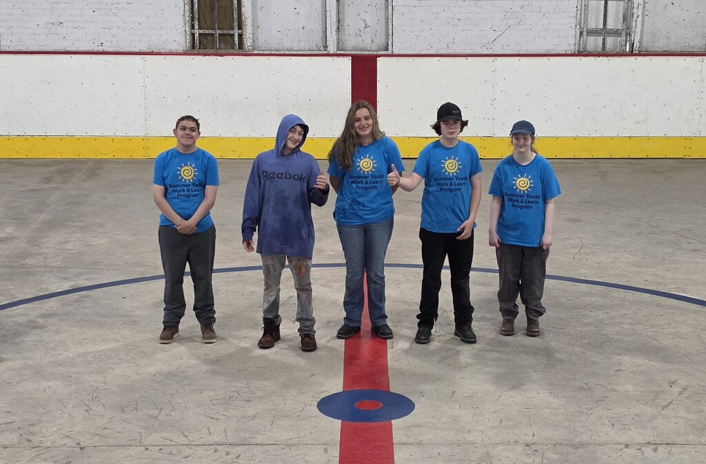 A group of students in bright blue Summer Youth Work & Learn t shirts pose for the camera on the freshly painted red center line of an ice hockey rink.