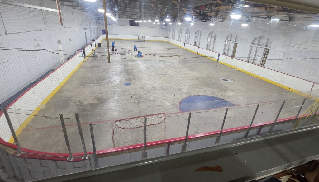 A view of an ice hockey rink. It is visible that the paint is fading. In the distance, students sit on the floor repainting the lines.