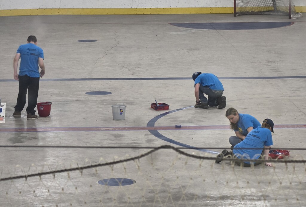 Students kneel and sit on the floor of an ice rink repainting the official lines on the concrete.