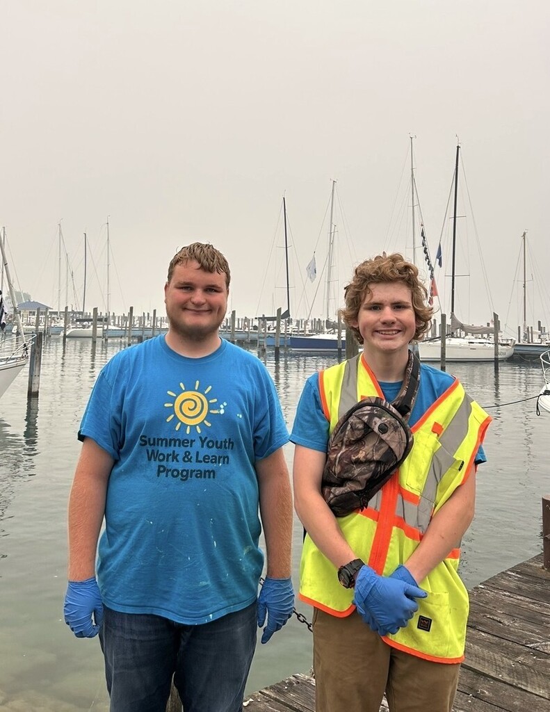 Two student workers on a dock smile for the camera. There are boats docked in the harbor behind them.