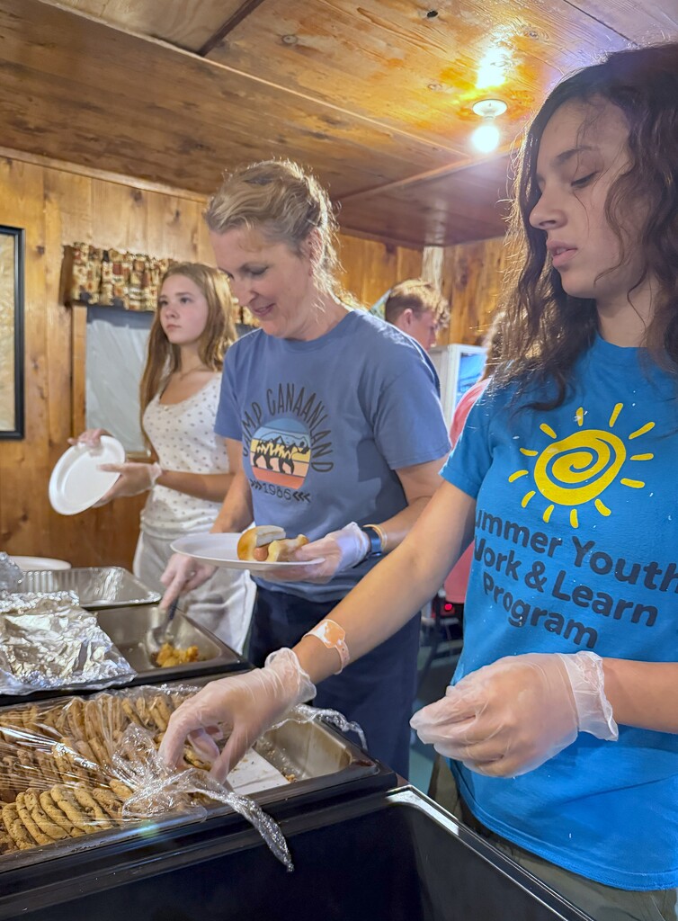 An adult and two students serve food in a lunch line.