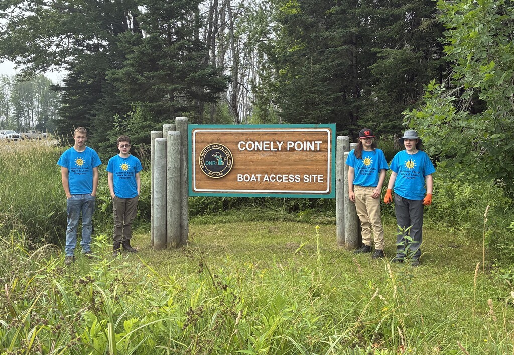 Two students in bright blue Student Youth Work & Learn stand on each side of a large wooden sign that says "Conely Point Boat Access. Site."