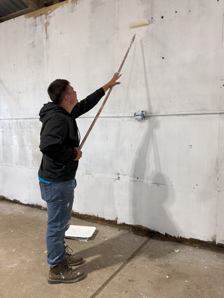 A young man uses a paint roller on a long handle to paint a wall white.