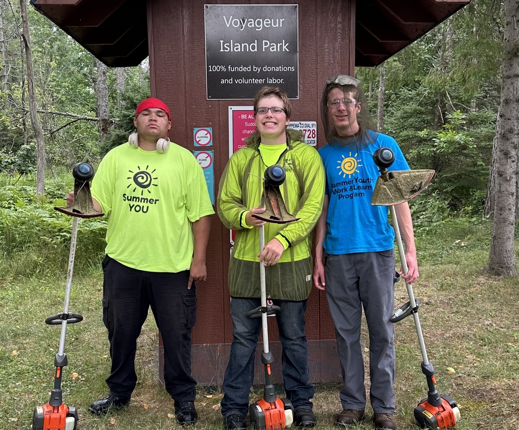 Three students pose with weed whips in front of a building with a sign that reads, "Voyageur Island Park. 100% funded by donations and volunteer labor. "