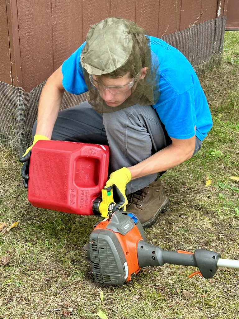 A student pours gas from a bright red can into a weed whip. He is wearing safety gloves and goggles. Mosquito netting covers his face.
