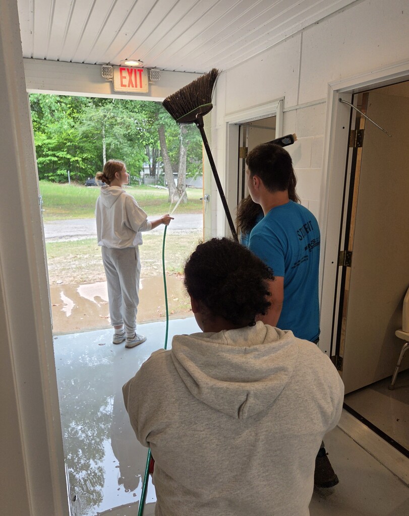 A student hoses down a concrete wall in a hallway of an open farigorunds building. 3 other students stand nearby with brooms.