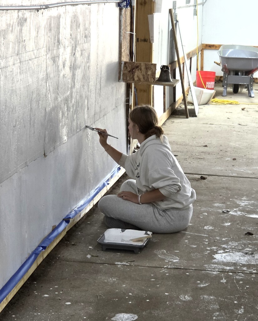 A student sitting on the floor, uses a paint brush to paint a wall white.
