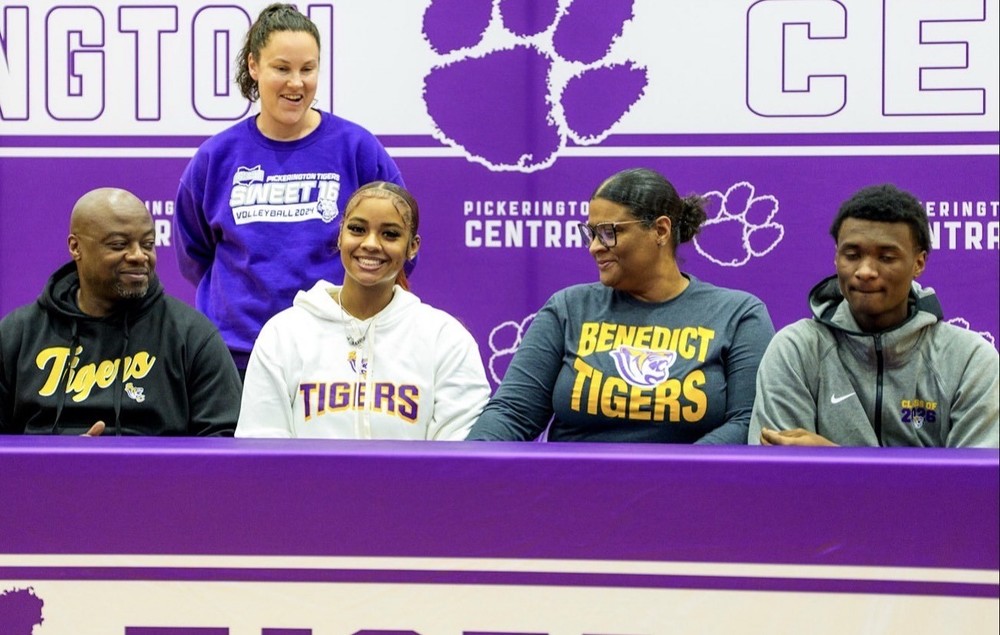 Moriah Smith sits at a signing table wearing a white “Tigers” hoodie and smiling, with three people seated beside her and a coach standing behind. Everyone is in front of a purple Pickerington Central backdrop featuring paw-print logos. Several individuals wear Benedict College or Tigers apparel, highlighting a college signing celebration.