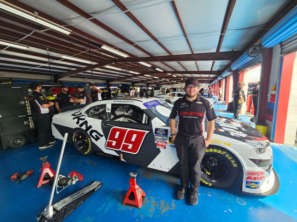 Aiden Malone, an Eastland-Fairfield Career & Technical Schools alumni, stands in front of a Viking Motorsports No. 99 NASCAR Xfinity Series car inside a race garage. He wears a black Viking Motorsports team shirt and hat, while other crew members work on cars in the background.