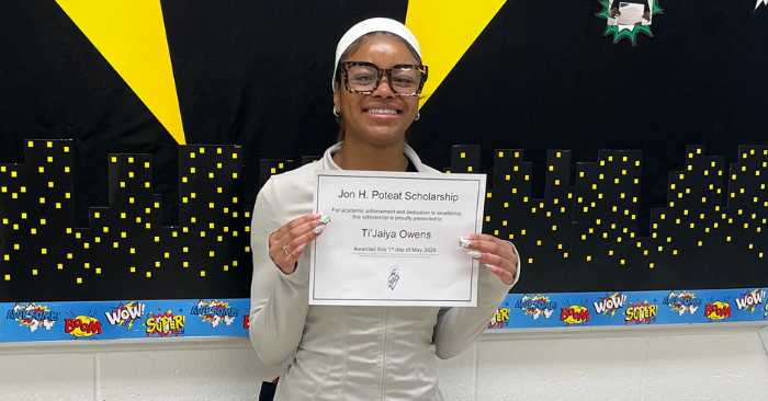 Student smiling while holding a Jon H. Poteat Scholarship certificate in front of a classroom display featuring a city skyline and superhero-themed decorations.