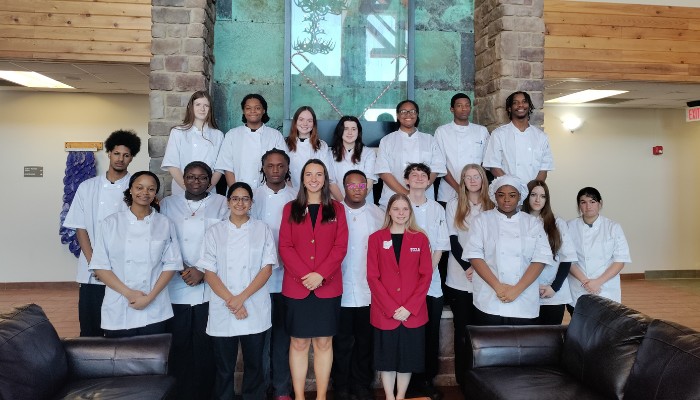 A group of culinary students wearing white chef coats pose for a photo indoors, standing in three rows behind two women in red blazers. The group is gathered in a lobby area with stone columns and a large window in the background, with couches visible in the foreground.