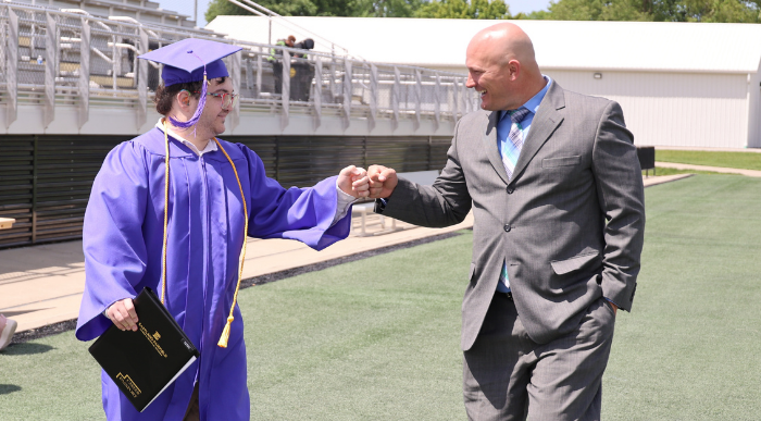 Graduate in a purple cap and gown fist bumps a staff member while holding a diploma cover during Eastland-Fairfield’s Senior Recognition ceremony at Fortress Obetz.