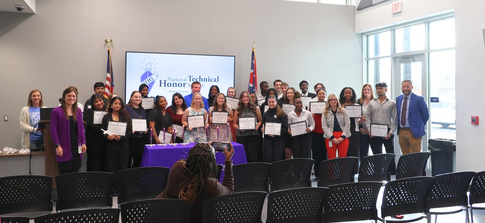 A group of Eastland Career Center students stand together holding certificates during the National Technical Honor Society induction ceremony, with staff members beside them and a presentation screen in the background.