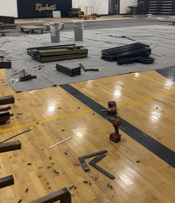 Interior of the EBF High School gymnasium during a bleacher replacement project. The gym floor is covered with protective tarps and construction materials, with sections of the old bleachers removed and stacked nearby. Tools and metal components are visible on the hardwood floor, while remaining bleachers line one wall. Basketball hoops, a scoreboard, and Rocket branding are visible in the background as preparations are underway for new ADA-compliant bleachers.