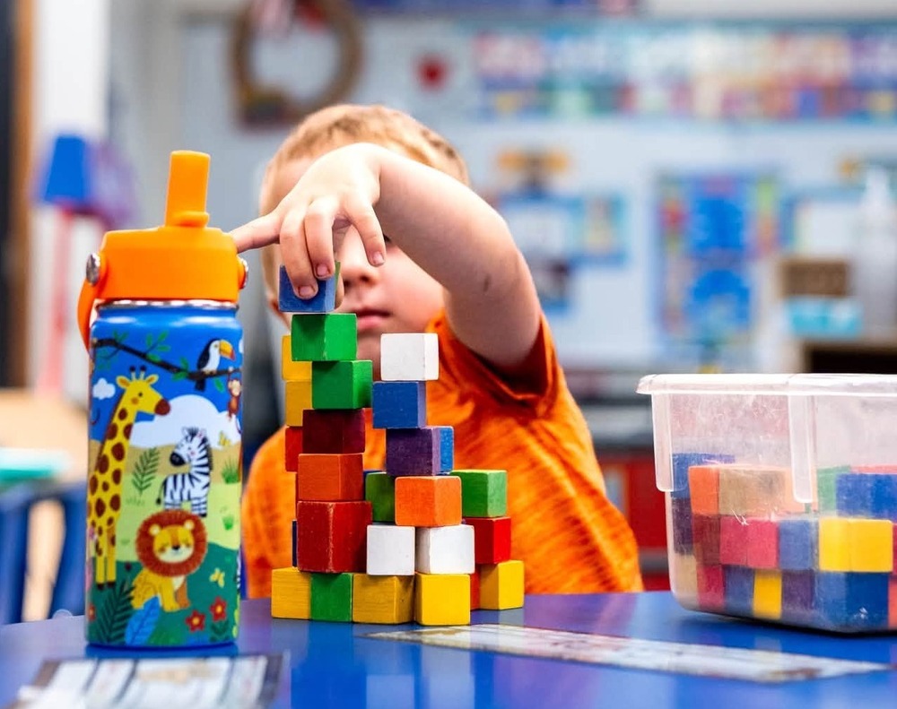 A preschool student stacks colorful wooden blocks at a blue classroom table. A colorful water bottle and a clear container of additional blocks are beside the block tower.