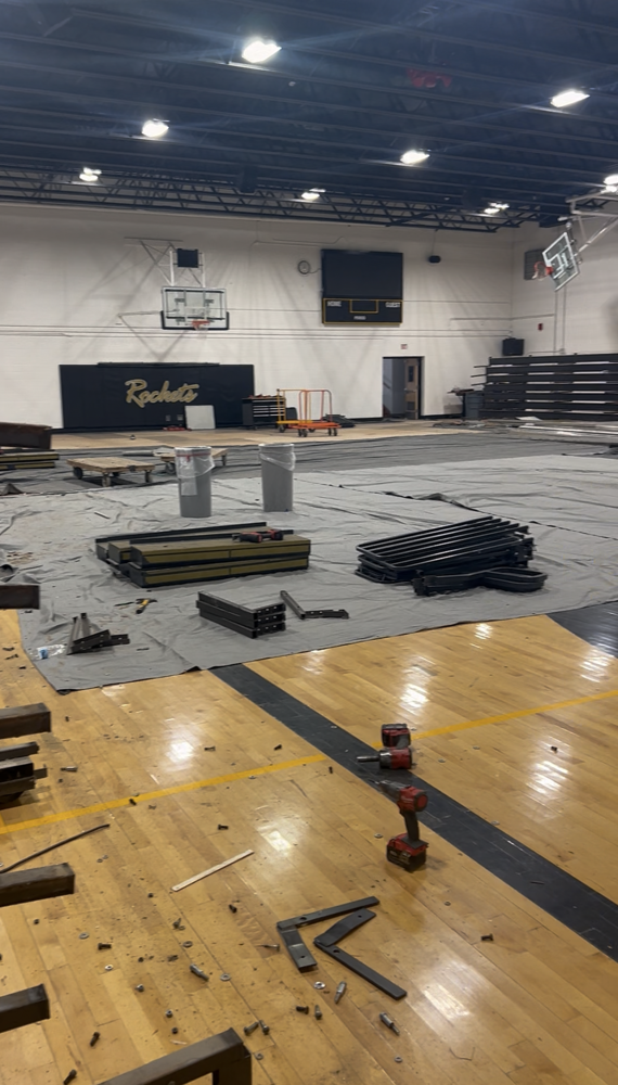 Interior of the EBF High School gymnasium during a bleacher replacement project. The gym floor is covered with protective tarps and construction materials, with sections of the old bleachers removed and stacked nearby. Tools and metal components are visible on the hardwood floor, while remaining bleachers line one wall. Basketball hoops, a scoreboard, and Rocket branding are visible in the background as preparations are underway for new ADA-compliant bleachers.