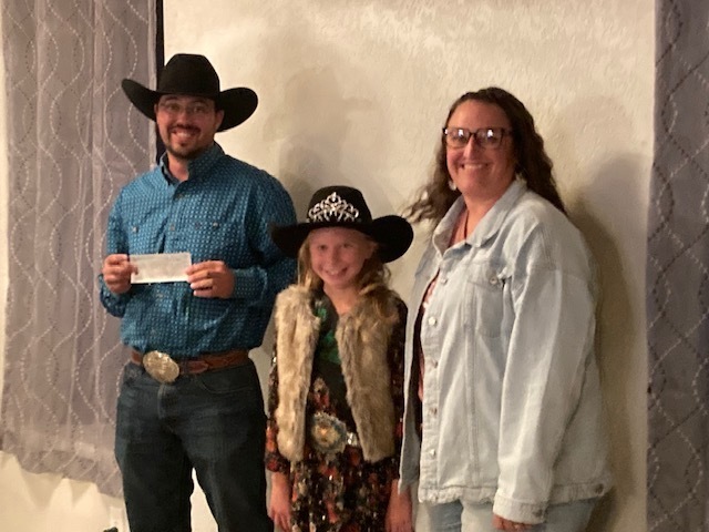 Man in cowboy hat holding check and smiling, standing next to a little girl in a cowboy hat and rodeo queen crown, and a woman wearing glasses and a denim jacket