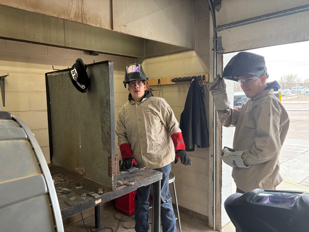 two boys in wearing safety coats, gloves, eye wear, and welding helmets smile at the camera