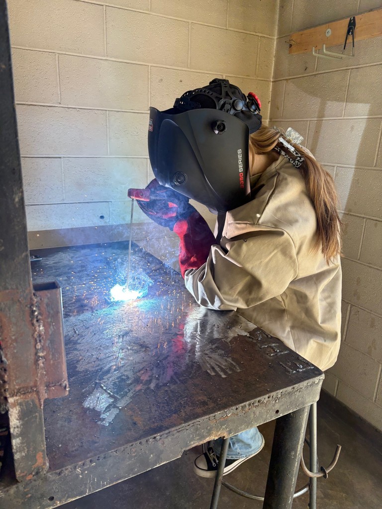 Girl in brown safety coat, gloves, and welding helmet welding with bright flash