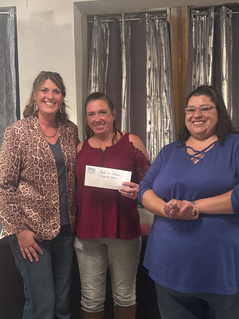 Three women stand smiling, holding an envelope with a donation for the Beef-to-School program.