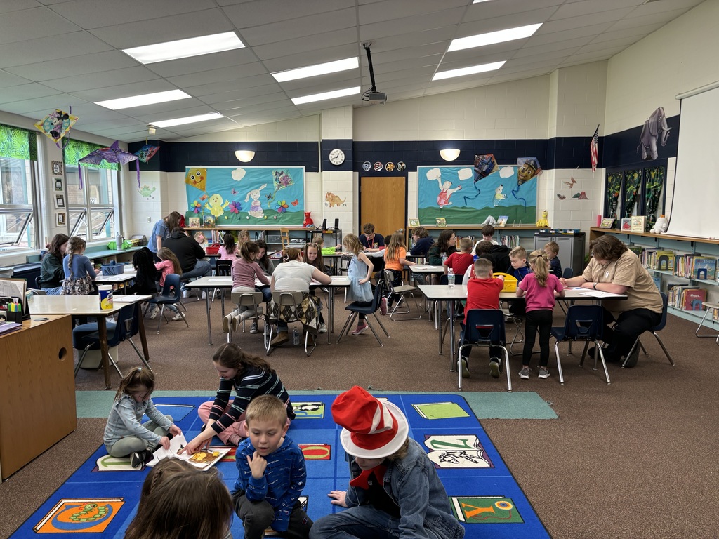 clasasroom view with studnets at tables and sitting on a bright colored carpet working with older students reading and coloring