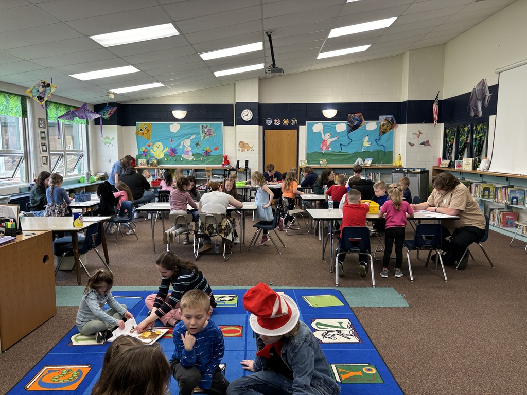 clasasroom view with studnets at tables and sitting on a bright colored carpet working with older students reading and coloring 