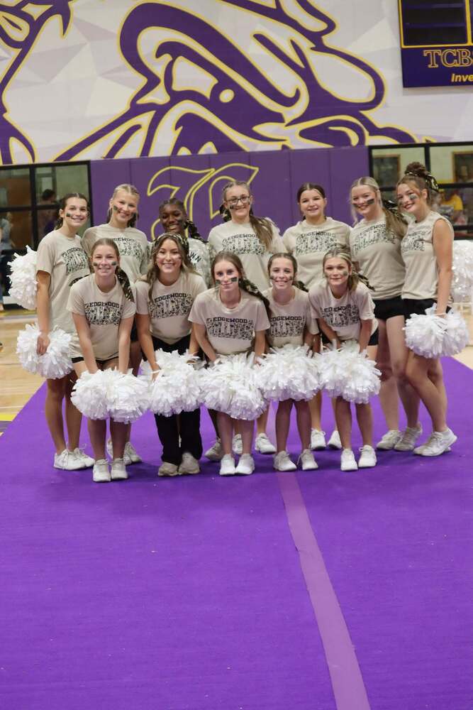Edgewood cheerleaders pose together during a pep rally in the school gym. The group wears matching Edgewood shirts with black shorts, white shoes, green face paint, and white pom-poms while standing on the purple gym floor in front of a large purple and gold Bulldog mural.