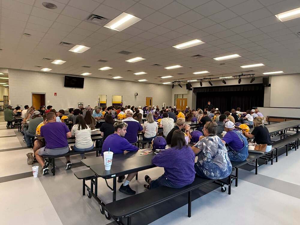 A large group of Edgewood Varsity Football players and their families gather in the school cafetorium for the annual Faith, Family, Football event. Families sit together at tables with purple and gold football helmets visible throughout the room while a speaker addresses the group from the front.