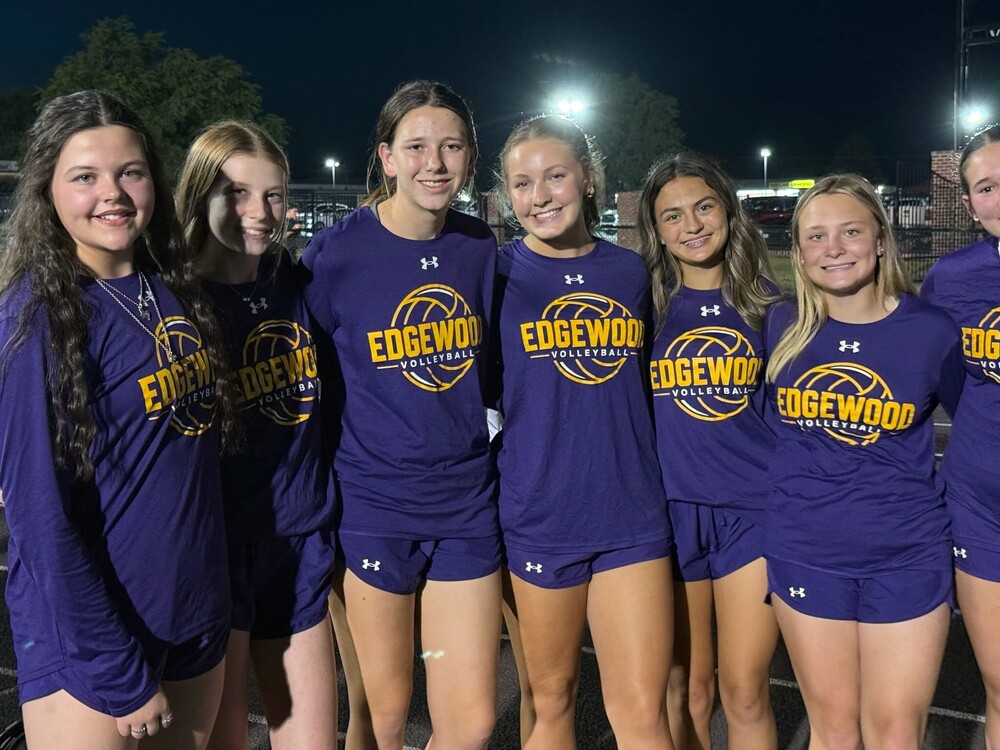 Edgewood volleyball players pose together outdoors at night wearing matching purple and gold Edgewood Volleyball shirts and purple athletic shorts. The group smiles for the camera under the stadium lights.