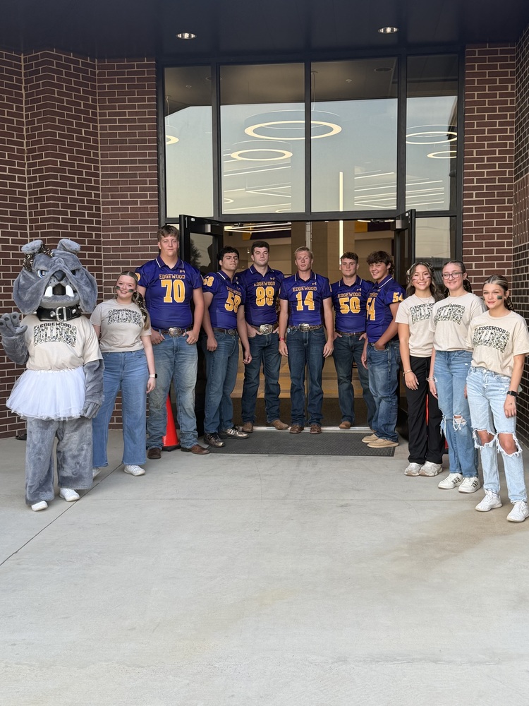 Edgewood High School varsity football players, cheerleaders, and the Bulldog mascot pose together outside a school entrance on game day. Football players wear purple and gold jerseys, while cheerleaders wear matching Edgewood shirts, capturing the school spirit and tradition of Friday mornings in Edgewood.