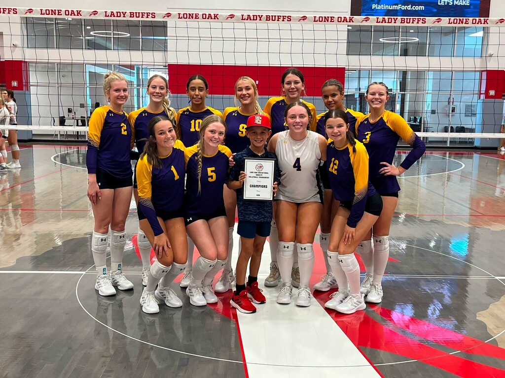 Edgewood High School varsity volleyball players pose together on the court at Lone Oak High School with a young supporter holding a Champions plaque from the Lone Oak Invitational Volleyball Tournament. The team is dressed in purple and gold uniforms and smiles together while celebrating their tournament championship.