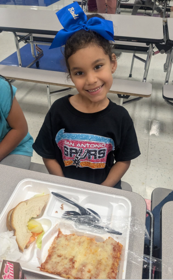 The picture shows a little girl smiling while eating lunch